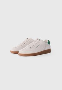 Baskets en daim crème avec un accent de couleur verte à l'arrière, semelle en caoutchouc marron et un design de laçage proéminent avec un détail de logo sur le côté.