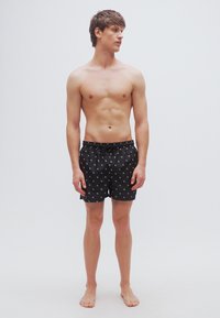 Μαύρα μαγιό σορτς με κορδόνι στη μέση, διακοσμημένα με ένα λευκό αφηρημένο σχέδιο. Φοριούνται από έναν άνδρα μοντέλο χωρίς πουκάμισο, όρθιο και ξυπόλυτο.
