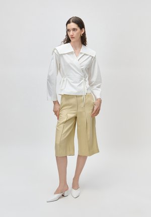 Donna che indossa una blusa avvolgente bianca con colletto ampio, pantaloni beige a gamba larga e corti alla caviglia, e scarpe bianche con tacco largo a punta, in piedi contro uno sfondo neutro.