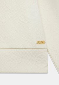Tissu de couleur crème avec un motif texturé. Il comporte une étiquette logo "GUESS" dorée sur l'ourlet, mettant en valeur le détail de la marque.