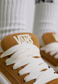 Brune suedette Vans-sneakers med hvite snører og logo på tungen, parret med hvite ribbestrikkede sokker. Nærbilde som viser tekstur og design.
