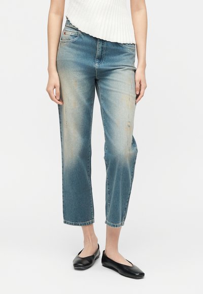Femme portant un jean bleu délavé raccourci et des ballerines noires, visible de la taille aux pieds sur un fond blanc uni.