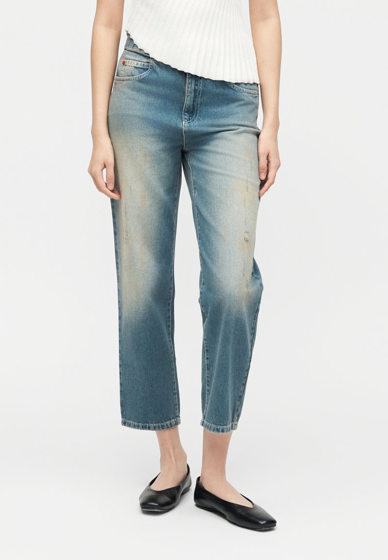 Γυναίκα που φοράει ξεθωριασμένο μπλε τζιν καμπάνα μέχρι τη μέση και μαύρα πέδιλα, εμφανιζόμενη από τη μέση μέχρι τα πόδια σε απλό λευκό φόντο.