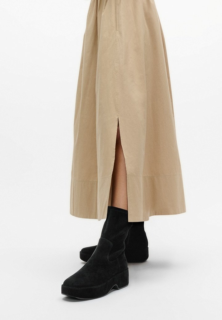 Zwarte enkellaarsjes van suede met een chunky zool, gedragen met een beige, lange rok met een zijsplit en een gladde textuur.