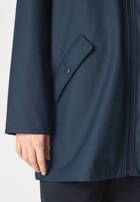 Marinblå vattentät jacka med en slät struktur, som har en dragkedjestängning, sidoficka med knapp och en avslappnad passform vid nederkanten.