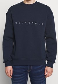 Námořnický modrý bavlněný svetr s bílým textem "ORIGINALS" napříč hrudí. Obsahuje kulatý výstřih a žebrované manžety a lem.