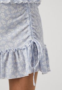 Gaiši zila svārku ar teksturētu ziedu rakstu, viļņotu apakšmalu un sānu rušingu. Iespējamas regulējamas kaklasaites. Kokvilnas maisījuma materiāls.