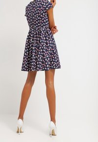 Kobieta w granatowej sukience z krótkim rękawem w motywy motyli, stojąca tyłem do kamery, w białych szpilkach.