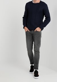 Marineblauer Strickpullover mit einem strukturierten Rautenmuster, graue Slim-Fit-Jeans und schwarze Sneakers mit roten Akzenten und weißen Sohlen.