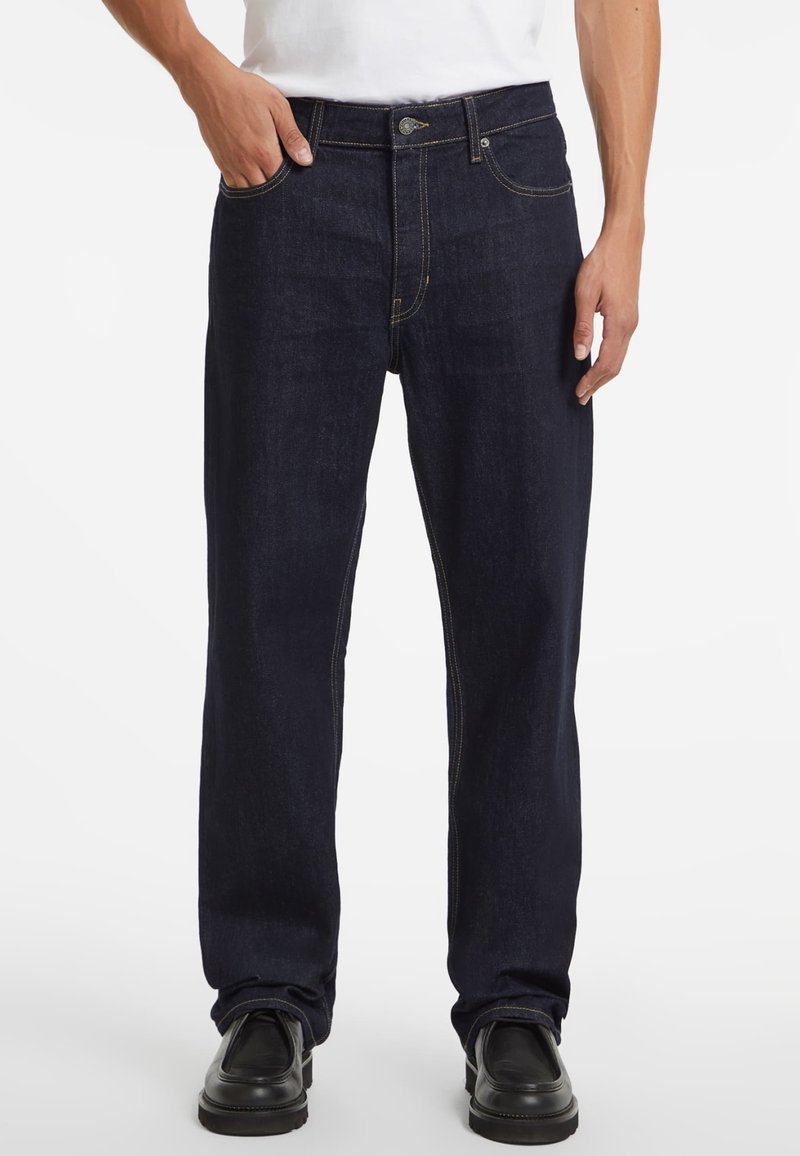 Jeans in denim blu navy con taglio dritto, cinque tasche, hardware in ottone e cuciture a contrasto. Indossati con una maglietta bianca e scarpe nere.