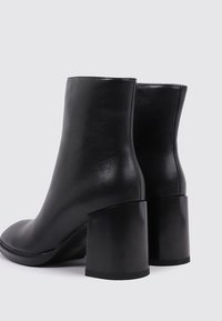 Botas de tobillo de cuero negro con una textura suave, punta redondeada y un tacón grueso, brillante y negro. Presenta un diseño minimalista y una silueta estilizada.