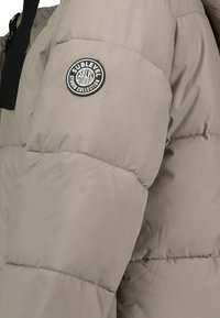 Beige Puffer-Jacke mit gestepptem Design und einem runden, schwarzen Logo-Patch am Ärmel. Mit durchgehendem Reißverschluss und hohem Kragen.