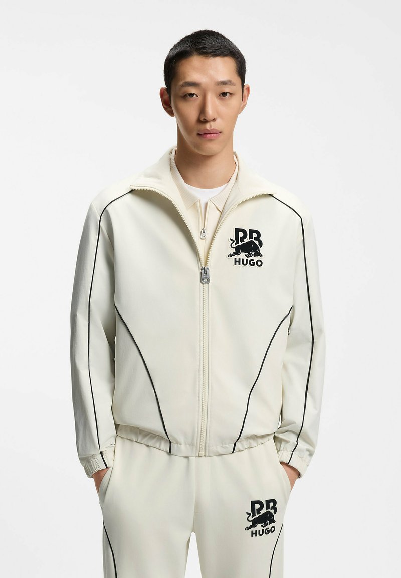 Jeune homme debout portant une veste blanche zippée et un pantalon avec des passepoils noirs et des logos de taureau noirs "RB HUGO" sur la poitrine et la cuisse.