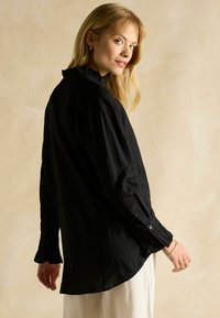 Woman with blonde hair wearing a loose black long-sleeve blouse with ruffled cuffs, paired with white pants, against a beige background.