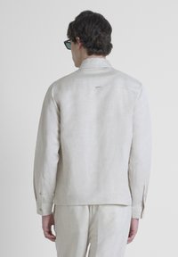Camicia di lino beige chiaro con maniche lunghe, colletto alla coreana e polsini con bottone. Presenta un piccolo logo vicino al collo e una consistenza morbida.