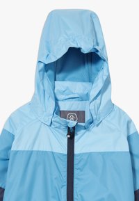 Světle modrá bunda s kapucí, s kontrastním tmavě modrým zipem a panelováním. Hladká textura, vhodná pro venkovní použití.