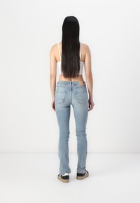 Jean en denim bleu skinny avec un délavage léger, comportant deux poches arrière et une étiquette en cuir beige à la taille. Le modèle porte des baskets noires et blanches.