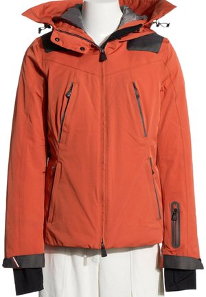 Chaqueta de invierno roja con cremallera, parches negros en los hombros, múltiples bolsillos con cremallera y capucha adjunta sobre un torso de maniquí blanco.