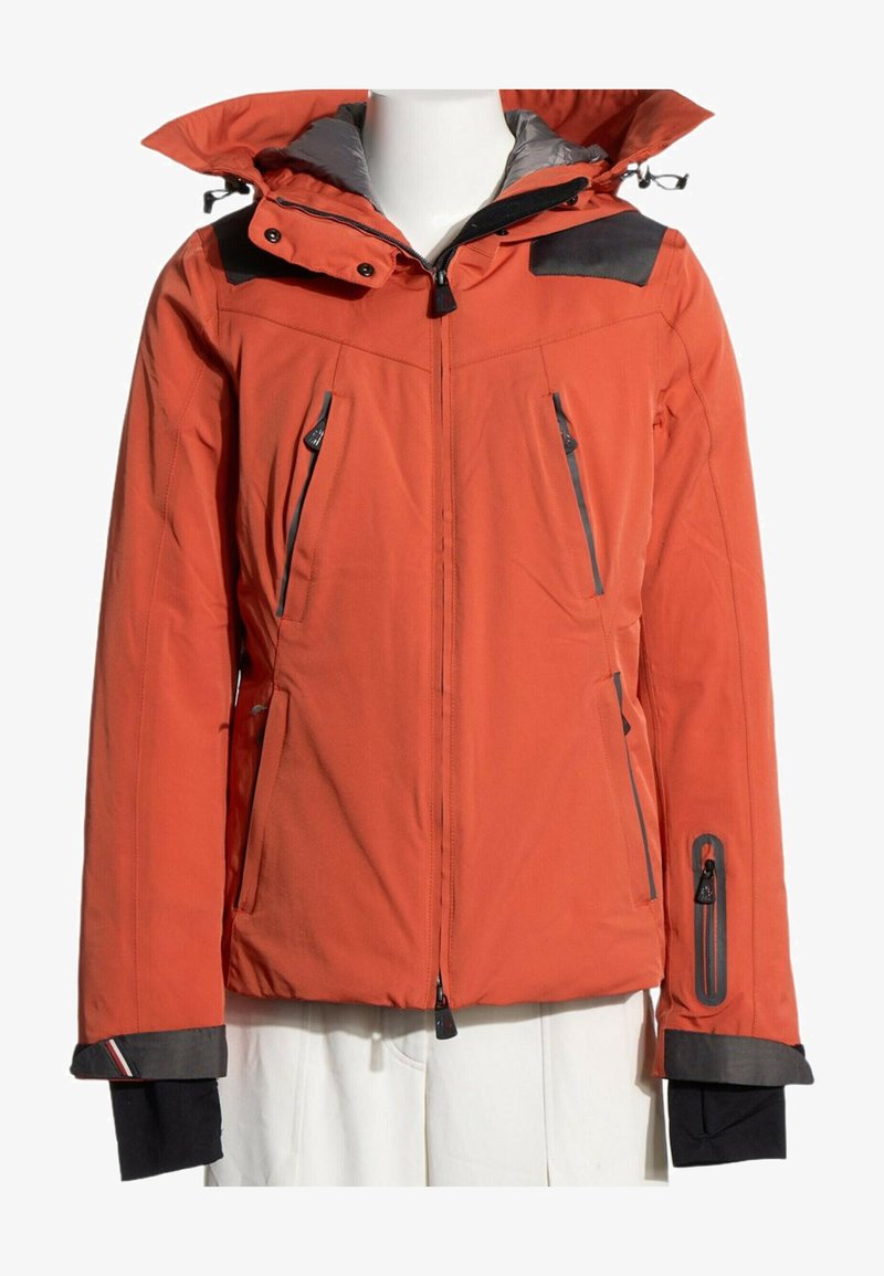 Veste d'hiver rouge zippée avec des empiècements noirs aux épaules, plusieurs poches zippées et une capuche fixée sur un torse de mannequin blanc.