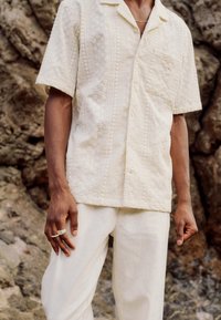 Homme portant une chemise crème texturée à manches courtes avec poche poitrine, pantalon crème et bagues argentées, debout devant un fond rocheux.