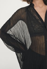 Schwarze durchsichtige Bluse mit plissierter Textur, offener Vorderseite und weitem Ärmeln. Mit einem dezenten Spitzenakzent darunter, der ein geschichtetes Design präsentiert.