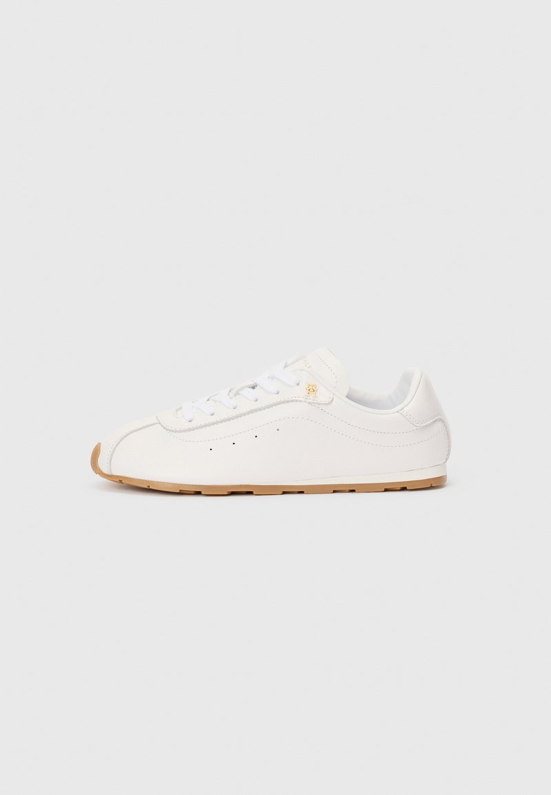 Χαμηλού προφίλ λευκό δερμάτινο αθλητικό παπούτσι με λευκές κορδόνες, μικρό χρυσό λογότυπο κοντά στις πάνω τρύπες και ανοιχτό καφέ λάστιχο.