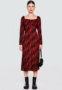 Kobieta w czerwono-czarną sukienkę midi w leopardzie, czarnych botkach sięgających kostek, długich kolczykach, czerwonej szmince i trzymająca czarną torebkę.