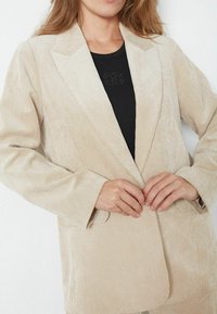 Béžový kordurový blejzer s vystúpenými klopami, prednými vreckami a textúrovanou látkou. Model si ho zapína cez čierny top.