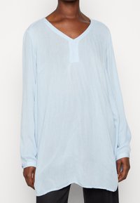 Person, die eine lässige, hellblaue Langarm-Tunika mit V-Ausschnitt und geknöpften Manschetten über einer dunklen Hose vor einem weißen Hintergrund trägt.