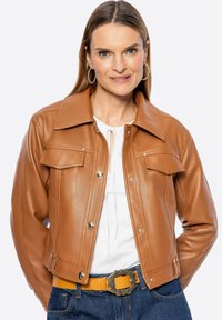 Giacca in pelle marrone con un design con colletto, bottoni a pressione frontali, due tasche sul petto e vita aderente. Indossata sopra una maglietta bianca e jeans blu.