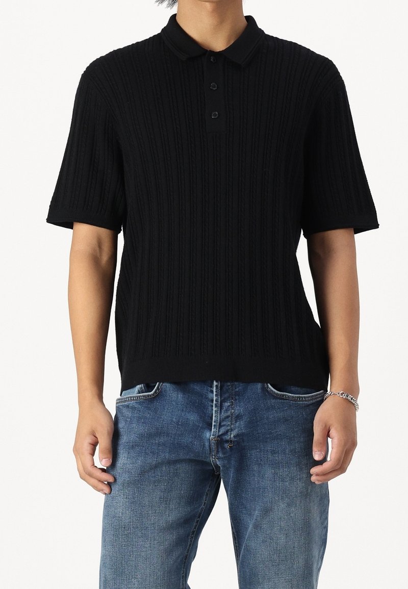 Camisa polo negra de manga corta con textura acanalada, que presenta un cuello clásico y una tapeta con tres botones, combinada con jeans azules.