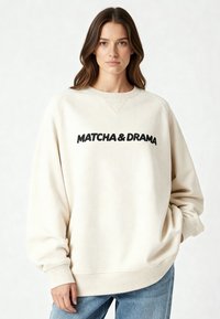 Sweat-shirt couleur crème avec texte noir "MATCHA & DRAMA". Comprend un col rond, des coutures tombantes aux épaules et des bracelets côtelés.