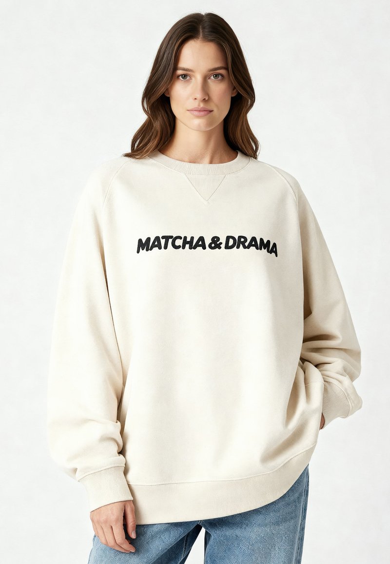 Sweat-shirt couleur crème avec texte noir "MATCHA & DRAMA". Comprend un col rond, des coutures tombantes aux épaules et des bracelets côtelés.