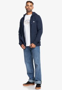 Námořnická modrá mikina na zip s předními kapsami, nošená přes bílý grafikou potisk t-shirt, v kombinaci se svetle modrými džíny a černými teniskami s hnědými podrážkami.