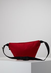 Red fabric fanny pack with a triangular shape, black adjustable strap, and buckle closure. Smooth texture, no visible patterns or accents.
