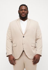 Beige suit jacket in smooth fabric with a single button closure and two front pockets. Styled with a white t-shirt underneath.