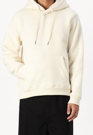 Cremefarbener Fleece-Hoodie mit strukturierter Oberfläche, Kapuze mit Kordelzug, Fronttasche sowie gerippten Bündchen und Saum. Darunter schwarze Hose.