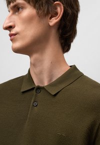 Olivgrünes Poloshirt aus strukturiertem Stoff, mit einem Kragen, zwei schwarzen Knöpfen und dezentem Branding auf der Brust.