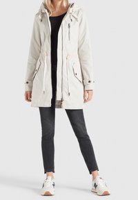 Parka a cappuccio beige chiaro con cordini regolabili, due tasche frontali e dettagli con zip. Abbinata a jeans scuri e sneaker bianche.