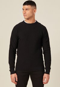 Schwarzer gerippter Strickpullover mit langen Ärmeln und Rundhalsausschnitt. Merkmale sind das strukturierte Material und die eng anliegenden Bündchen. Getragen mit einem weißen Unterhemd.
