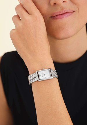 Zilveren rechthoekig polshorloge met een witte wijzerplaat, stijl van een mesh-armband, gedragen op een lichtgekleurde pols. Zwarte achtergrond.
