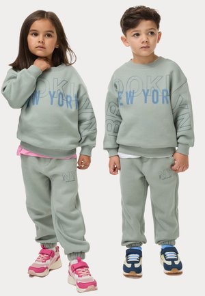Lichtgroene sweaters met de tekst "BROOKLYN" en "NY", gecombineerd met bijpassende sweatpants. Kleurrijke sneakers: roze aan de linkerkant, blauw aan de rechterkant.