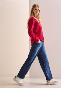 Chemise rouge à manches longues, jean bleu ample et baskets blanches avec une touche de rouge, se tenant devant un fond neutre.