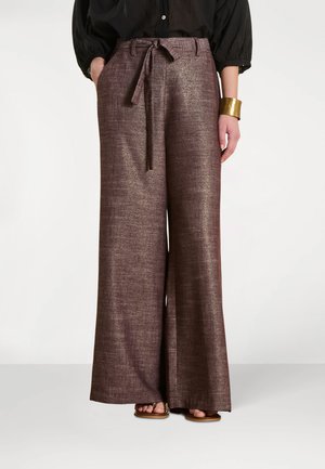 Pantalon marron à jambes larges et taille haute avec ceinture en tissu nouée à la taille, porté avec un chemisier noir, un bracelet manchette doré et des sandales marron.