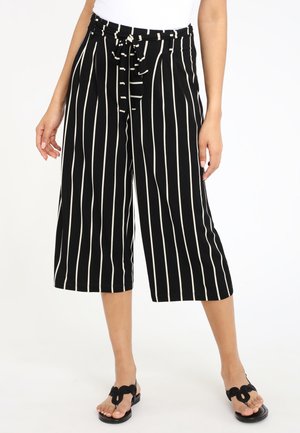 Donna che indossa pantaloni neri a gamba larga e corta con righe verticali bianche e cintura annodata in vita, abbinati a sandali neri.