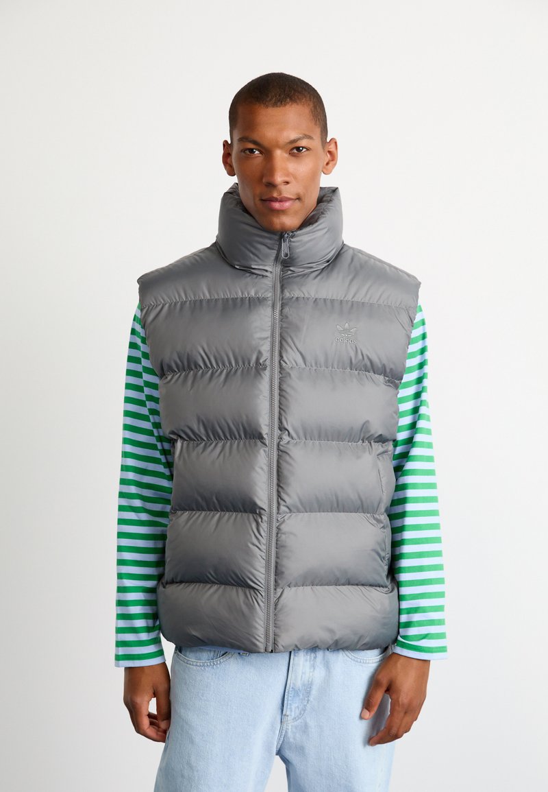 Grå puffervest med høj krave og lynlås, med quiltet design. Båret over en grøn og hvid stribet langærmet skjorte.