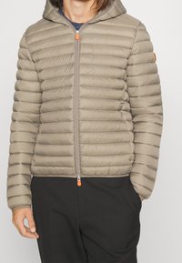 Beige gewatteerd jack met capuchon, voorzien van een horizontaal doorgestikt ontwerp, een ritssluiting aan de voorkant met een oranje trekker, en een slanke pasvorm. Geschikt voor vrijetijdsgebruik.