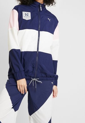 Persoon draagt een marineblauw, wit en roze trainingsjack en bijpassende broek met Puma-logo's en een "92 EST" patch op het jack.