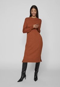 Rozsdagyűrű színű, hosszú ujjú kötött ruha oldalsó sliccekkel és kerek nyakkivágással, fekete térdig érő csizmával párosítva. Sima textúra, testhezálló sziluett.