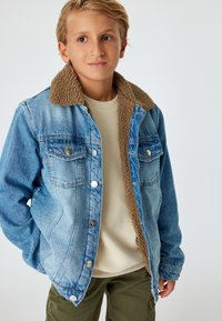 Jeansjacka med en brun fårskinnskrage, två bröstfickor och silverdetaljer. Bärs över en ljusbeige tröja, tillsammans med gröna byxor.